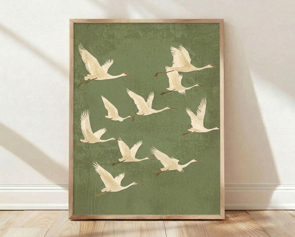 May include: Framed poster with olive green background featuring nine cream-colored cranes in flight, displayed in light wooden frame leaning against white wall on light wood floor with soft natural lighting and fabric shadows