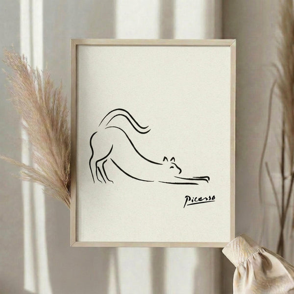 May include: Minimalist line drawing of a stretching cat in black ink on cream textured paper, displayed in light wood frame. Styled with dried pampas grass and neutral fabric against soft beige curtains in natural lighting. Simple continuous line art shows cat in downward stretch pose with curved tail and pointed ears.