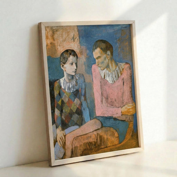 May include: Framed reproduction of Picasso's 1905 painting showing two young acrobats in harlequin costumes sitting together. One wears a diamond-patterned outfit in blues and earth tones, the other in a pink top. The artwork features muted blue and ochre backgrounds with the artist's signature Rose Period style. Light wooden frame leans against white wall.