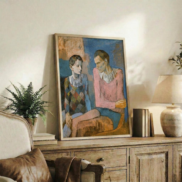 May include: Framed reproduction of Picasso's 1905 painting showing two seated figures - a young person in a diamond-patterned harlequin costume and an adult in a pink shirt. The artwork features muted blues, pinks, and earth tones in Picasso's early period style. Displayed on a rustic wooden sideboard alongside books, a ceramic vase, and potted fern in a modern living room with cream walls and neutral furnishings.