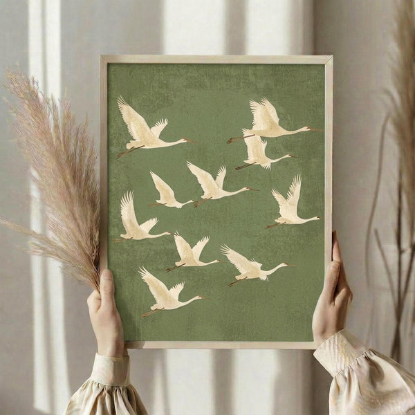 May include: Person holding framed artwork featuring nine cream-colored cranes in flight against sage green textured background, shown with dried pampas grass in minimalist neutral setting with natural lighting and flowing curtains