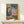 May include: Framed reproduction of Picasso's 1905 painting showing two seated acrobats in harlequin costumes against blue background, displayed on light wood console table beside white wrapped gift with small evergreen sprig, set against cream wall in minimalist interior setting.