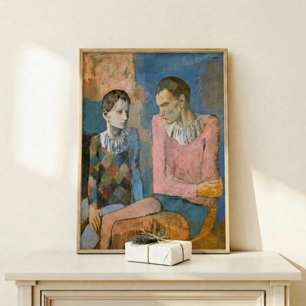 May include: Framed reproduction of Picasso's 1905 painting showing two seated acrobats in harlequin costumes against blue background, displayed on light wood console table beside white wrapped gift with small evergreen sprig, set against cream wall in minimalist interior setting.