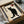 May include: Framed artwork featuring a black cat silhouette with golden eyes sitting against an aged cream background, accompanied by Japanese kanji characters on the left side and a red heart symbol with black text above. The piece is displayed in a black frame within an open cardboard shipping box surrounded by white tissue paper, with wooden dowels visible nearby.
