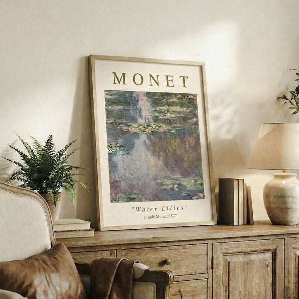 May include: Framed Monet Water Lilies poster print displayed on rustic wooden sideboard in warm-toned living room setting. Print shows impressionist pond scene with floating lily pads in soft blues, greens, and purples. Natural wood frame complements weathered cabinet, potted fern, ceramic vases, and stacked books. Cozy neutral decor with textured throw pillow visible.