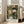 May include: Framed botanical art print leaning against white wall beside beige armchair in bright living room. Print features intricate floral pattern with pink, white, and cream flowers among detailed green foliage on dark background. Light wood frame with white mat displays artist attribution text below artwork. Natural sunlight streams through tall windows onto hardwood floors.
