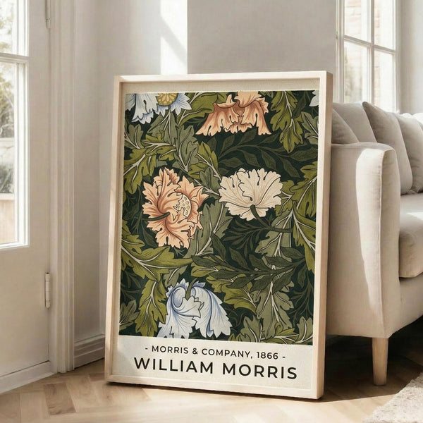 May include: Framed botanical art print leaning against white wall beside beige armchair in bright living room. Print features intricate floral pattern with pink, white, and cream flowers among detailed green foliage on dark background. Light wood frame with white mat displays artist attribution text below artwork. Natural sunlight streams through tall windows onto hardwood floors.