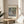 May include: Framed art print featuring Monet's impressionist water lilies painting with soft blues, greens, and pink reflections on water surface. White matted poster with "MONET" text and artwork details displayed on round wooden side table next to beige upholstered chair with throw pillow. Natural sunlight creates shadows on cream wall, with plant leaves visible in corner of bright, minimalist living room setting.