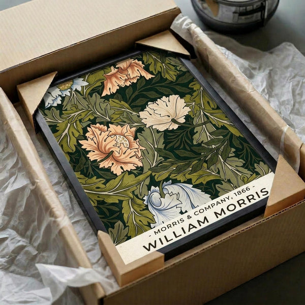 May include: Framed botanical art print featuring William Morris-style design with ornate green foliage and coral-colored flowers on dark background, displayed in black frame inside open cardboard shipping box with protective wrapping paper on white surface