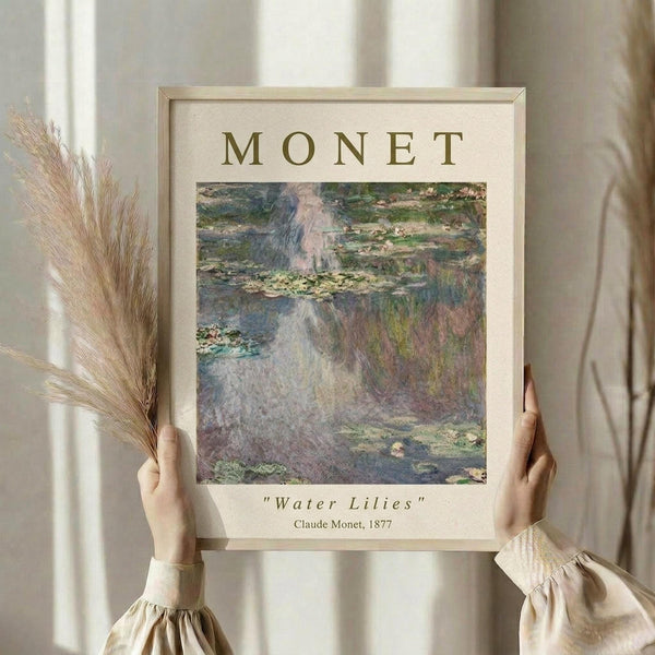 May include: Hands holding a framed art print of Monet's Water Lilies painting showing impressionist pond scene with floating lily pads in soft blues, greens, and pinks. Light wood frame with cream matting displays "MONET" at top and artwork details below. Person wears beige ruffled sleeves, holds dried pampas grass stems, standing against neutral curtained background with soft natural lighting.