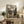 May include: Framed anime poster displayed on rustic wooden sideboard in cozy living room setting. Poster shows two samurai characters dueling with swords in traditional Japanese village scene, featuring muted earth tones and vintage-style artwork. Scene includes white armchair, potted fern, ceramic vases, stacked books, and warm ambient lighting on cream walls.