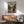 May include: Framed anime poster showing two sword-fighting characters in traditional Japanese village setting, displayed above wooden sideboard with white ceramic lamp in modern living room. Mid-century green upholstered chair with wooden arms sits nearby. Natural sunlight creates geometric shadows on white wall. Poster features muted earth tones and vintage film poster styling.