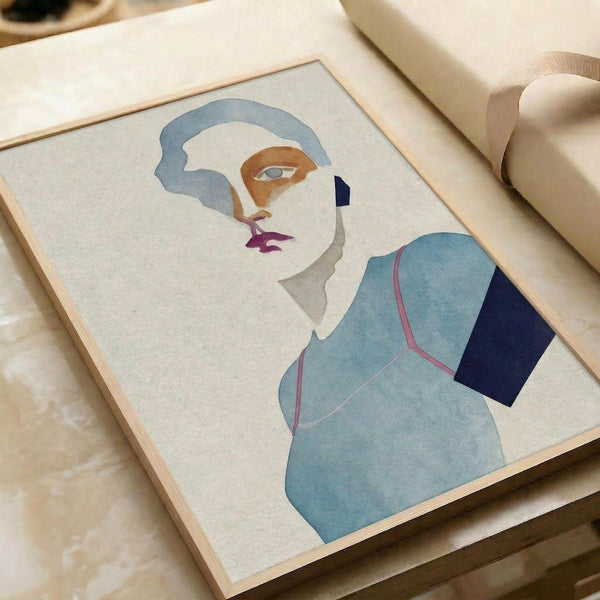 Framed minimalist portrait artwork in muted blue, orange, and purple tones showing an abstract female figure with stylized facial features and geometric clothing shapes. The print sits on a light wooden surface next to rolled paper tubes, displaying soft watercolor-like textures within clean geometric forms against a cream background.