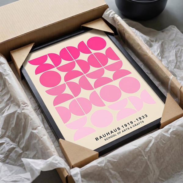 May include: Framed geometric art print with pink gradient circles and leaf shapes on cream background, displayed in black frame emerging from brown cardboard packaging with white tissue paper, alongside a wooden rolling pin and black bowl on neutral surface.
