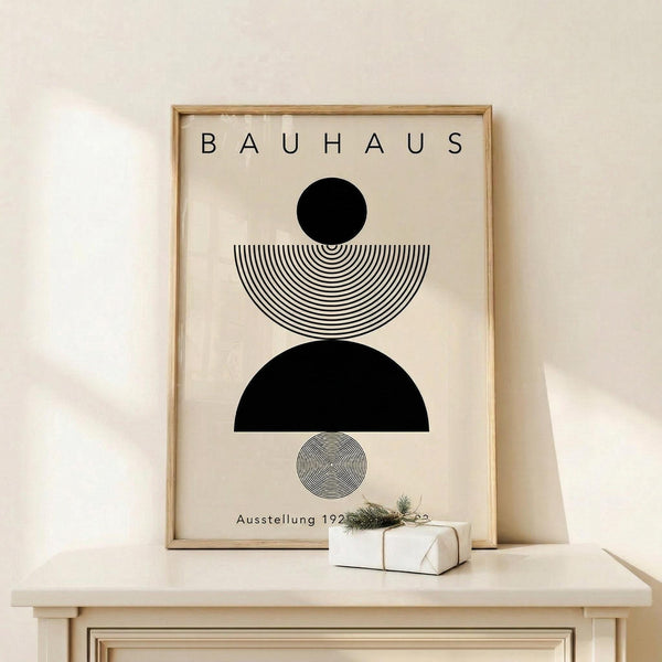 May include: Framed Bauhaus-style poster with geometric black and white design featuring circles and semicircles with concentric line patterns, displayed on light wood surface against cream wall. Small wrapped gift with evergreen sprig sits nearby. Modern minimalist home decor in neutral tones.