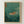 May include: Framed vintage-style Japanese woodblock print featuring an orange and black striped tiger swimming through turquoise-green churning waves. Red text cartouche in upper left corner contains Japanese characters. Print shows weathered, aged texture with visible wear marks. Natural wood frame hangs on plain white wall. Traditional ukiyo-e art style with flowing water patterns and small vegetation details in corners.