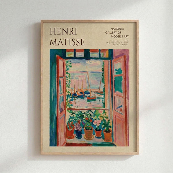 May include: Framed vintage museum poster featuring Henri Matisse's colorful fauve painting of an open window with potted plants on the sill overlooking boats in a harbor. The artwork shows vibrant blues, greens, oranges and pinks in Matisse's distinctive bold style. Natural wood frame mounted on white wall in modern interior setting.
