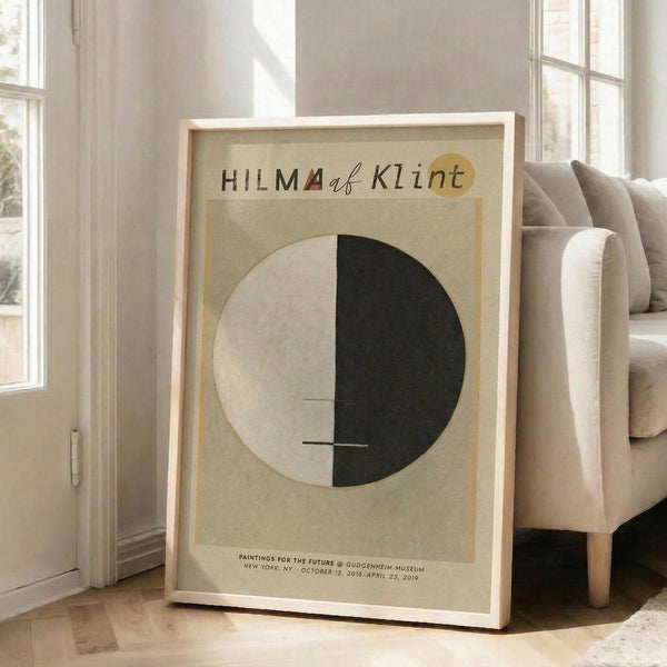 May include: Framed abstract art poster leaning against white wall next to beige upholstered chair. Cream-colored print features circular design split between white and dark green halves with thin horizontal lines crossing through center. Text at top reads "HILMA af Klint" with small red triangle accent. Exhibition details appear at bottom in small print. Natural wood frame, bright sunlit room with large windows.