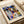 May include: Abstract geometric artwork print in wooden frame featuring colorful overlapping shapes in blues, greens, oranges, and purples. Text reads "PAUL KLEE" at top and "BAUHAUS ARCHIV" and "1919-2019 JUBILÄUM" at bottom. Frame sits on white bedding next to rolled paper, showing the modernist composition with triangular and rectangular forms creating a complex layered pattern.