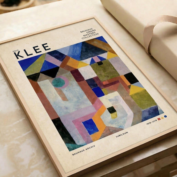 May include: Abstract geometric artwork print in wooden frame featuring colorful overlapping shapes in blues, greens, oranges, and purples. Text reads "PAUL KLEE" at top and "BAUHAUS ARCHIV" and "1919-2019 JUBILÄUM" at bottom. Frame sits on white bedding next to rolled paper, showing the modernist composition with triangular and rectangular forms creating a complex layered pattern.