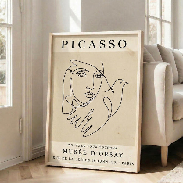May include: A framed beige poster with black line art showing a woman's face and hands holding a dove, displayed on light wooden floors against white walls next to a cream-colored armchair. The minimalist artwork features "PICASSO" at the top and "MUSÉE D'ORSAY" with French text below. Natural sunlight streams through tall windows, creating soft shadows on the neutral-toned interior.