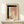 May include: Abstract watercolor artwork in wooden frame displaying black brushstrokes forming geometric shapes with soft pink and cream washes, dotted vertical line detail, displayed on light wood shelf alongside small wrapped gift with evergreen sprig against cream wall