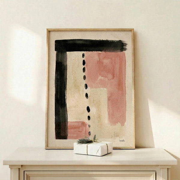 May include: Abstract watercolor artwork in wooden frame displaying black brushstrokes forming geometric shapes with soft pink and cream washes, dotted vertical line detail, displayed on light wood shelf alongside small wrapped gift with evergreen sprig against cream wall
