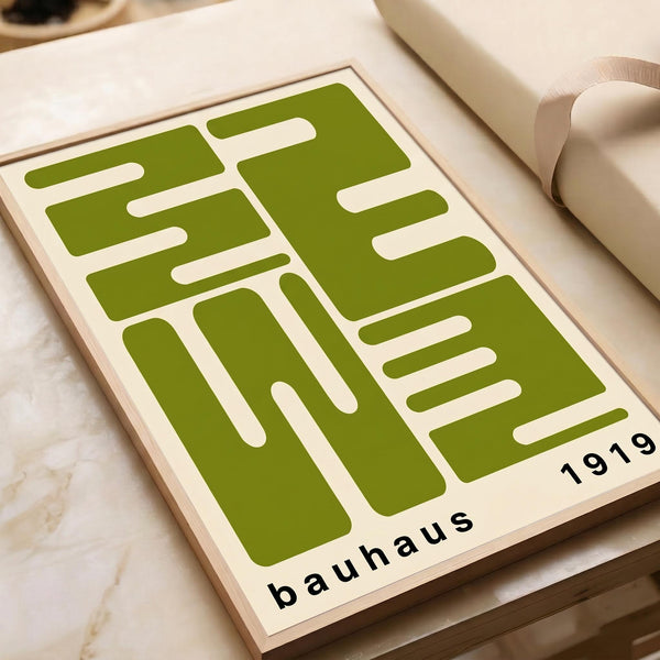 May include: Green and cream geometric abstract art print displayed on a wooden surface next to rolled paper tubes. The minimalist design features bold angular shapes and lines in olive green against a light background, with "bauhaus 1919" text at the bottom. The print appears to be on white paper with clean modern styling.