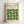 May include: Framed poster with cream background displaying "BAUHAUS" text and geometric green leaf-like shapes arranged in a grid pattern. Light wood frame leans against white wall with soft natural lighting casting shadows.