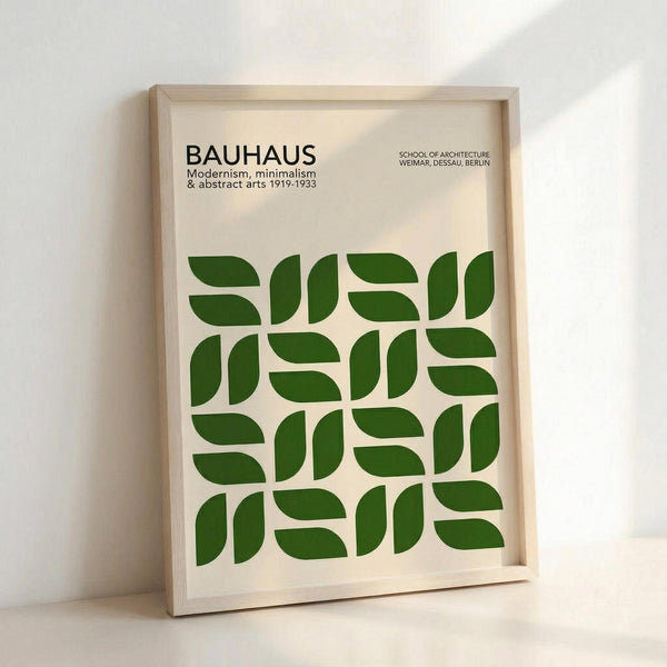 May include: Framed poster with cream background displaying "BAUHAUS" text and geometric green leaf-like shapes arranged in a grid pattern. Light wood frame leans against white wall with soft natural lighting casting shadows.
