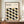May include: Framed Bauhaus-inspired poster with geometric leaf pattern in black, olive green, and taupe on cream background, displayed on wooden sideboard beside white ceramic vase and stacked books in neutral-toned room with white paneled walls.