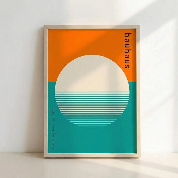 A framed poster featuring a minimalist geometric design with a large white circle representing the sun against a bright orange sky above teal water. Horizontal white lines create a reflection effect across the lower half of the circle. The word "bauhaus" appears vertically in black text on the right side, with "Ausstellung Juli Sept. 1923" in small text along the left margin. The artwork is displayed in a light wood frame against a white wall.