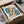 May include: Framed Bauhaus-style poster with geometric design featuring teal numbers "010", orange and black abstract shapes, and concentric square patterns on cream background. The black-framed print is nestled in cardboard packaging with white tissue paper, sitting on a neutral surface next to what appears to be a dark ceramic object.