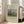 May include: Framed art print of Henri Rousseau's pastoral painting showing cows grazing in a green meadow with tall poplar trees and a large rounded tree, two small figures in the scene. Cream-colored poster with museum-style text layout displayed in light wooden frame, leaning against white wall next to beige upholstered chair in bright, airy room with large windows.