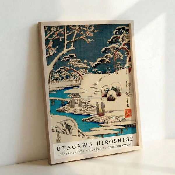 May include: Framed Japanese woodblock print reproduction showing a snow-covered winter garden scene with traditional stone lantern, curved bridge over blue stream, snow-laden pine trees, and small figures in traditional dress. Artwork features classic ukiyo-e style with muted blues, whites, and earth tones, displayed in light wooden frame against white wall.