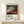 May include: Framed art print featuring Hokusai's iconic Mount Fuji woodblock print with reddish-brown mountain against blue sky with white clouds, displayed on light wood console table beside small wrapped white gift box with evergreen sprig, against cream wall with natural lighting