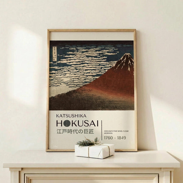 May include: Framed art print featuring Hokusai's iconic Mount Fuji woodblock print with reddish-brown mountain against blue sky with white clouds, displayed on light wood console table beside small wrapped white gift box with evergreen sprig, against cream wall with natural lighting