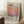 May include: Framed art print leaning against white wall beside wooden side table with coffee cup. Print features gradient sunset colors in orange, pink, and purple with "Mark Rothko" text and exhibition details. Light wood frame, sitting on white textured rug next to beige fabric chair in bright, naturally lit room.