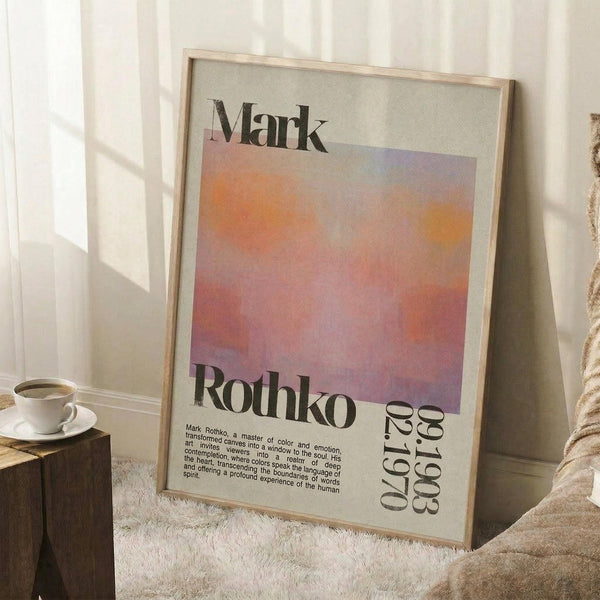 May include: Framed art print leaning against white wall beside wooden side table with coffee cup. Print features gradient sunset colors in orange, pink, and purple with "Mark Rothko" text and exhibition details. Light wood frame, sitting on white textured rug next to beige fabric chair in bright, naturally lit room.