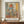 May include: Abstract painting poster in wooden frame displayed on walnut credenza beside white ceramic vase and books. Artwork features geometric shapes in muted orange, blue, coral and beige tones with soft, layered brushstrokes. Room has cream walls with white wainscoting and warm natural lighting.