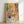 May include: Framed art print in light wood frame leaning against white wall on light wooden floor. Artwork features colorful Matisse-inspired interior scene with potted plants, patterned chair, decorative tiles, and "MATISSE" text at bottom. Vibrant yellows, pinks, greens, and blues with botanical and geometric patterns throughout.