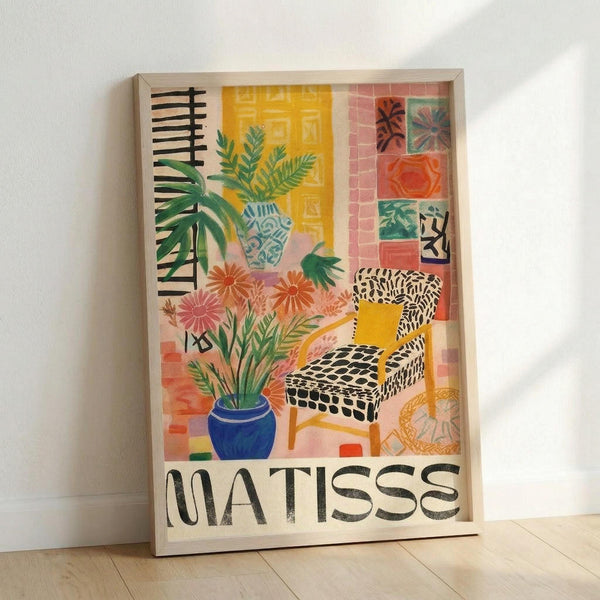 May include: Framed art print in light wood frame leaning against white wall on light wooden floor. Artwork features colorful Matisse-inspired interior scene with potted plants, patterned chair, decorative tiles, and "MATISSE" text at bottom. Vibrant yellows, pinks, greens, and blues with botanical and geometric patterns throughout.