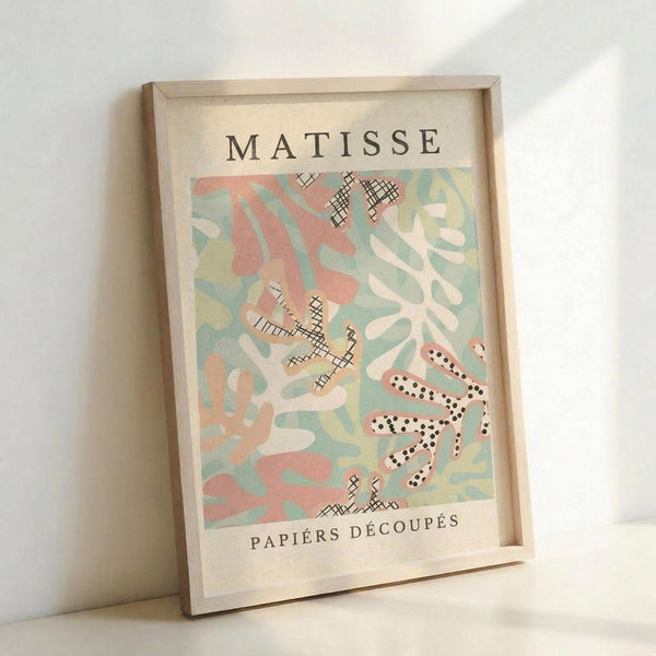 May include: Framed vintage Matisse exhibition poster with abstract cut-out shapes in coral pink, sage green, cream and yellow, featuring black cross-hatched patterns and dotted elements. Light wood frame leans against white wall, casting soft shadows. The artwork shows organic, flowing forms reminiscent of paper cutouts with "MATISSE" and "PAPIERS DÉCOUPÉS" text at top and bottom.