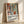 May include: Framed poster with Matisse-inspired artwork showing colorful still life scene with vases, plants, and geometric shapes in terracotta, green, and blue tones. Light wood frame rests against white wall with natural shadows. Text below artwork reads "Celebrating Colors of Life" with descriptive paragraph about adding color to spaces.
