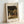 May include: Framed art print featuring a woman in white dress with parasol walking through a lush garden with flowering trees and foliage in warm earth tones. The print has "CLAUDE MONET" at top and "EXHIBITION ART" at bottom on cream-colored paper within a light wood frame, leaning against a white wall.