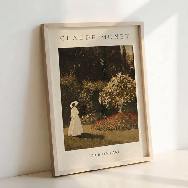 May include: Framed art print featuring a woman in white dress with parasol walking through a lush garden with flowering trees and foliage in warm earth tones. The print has "CLAUDE MONET" at top and "EXHIBITION ART" at bottom on cream-colored paper within a light wood frame, leaning against a white wall.