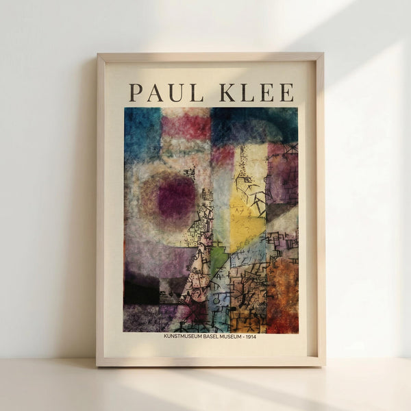 May include: Framed Paul Klee museum exhibition poster displayed against white wall with natural lighting creating soft shadows. The abstract artwork features vibrant watercolor-style patches in blues, purples, yellows, and earth tones with geometric line drawings and architectural elements overlaid. Light wood frame complements the cream-colored mat border. Text reads "KUNSTMUSEUM BASEL MUSEUM - 1914" below the colorful modernist composition.