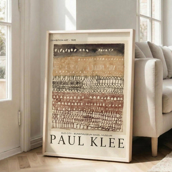 May include: Framed exhibition poster featuring Paul Klee's abstract geometric artwork with earth-toned horizontal bands of triangles, dots, and linear patterns in browns, creams, and dark tones. Light wooden frame leaning against white wall next to beige upholstered sofa in bright, sunlit room with hardwood floors and tall windows.