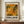 May include: Abstract art exhibition poster in brown frame leaning against white paneled wall on wooden credenza. Poster features vibrant orange, yellow, and green paint splatters with black text reading "POLLOCK" and "EMBRACE THE CHAOTIC REALM OF CREATIVITY" with exhibition dates. White ceramic vase and stacked books sit nearby on the warm wood surface.