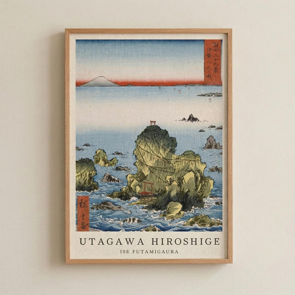 May include: Framed Japanese woodblock print reproduction mounted on white wall, featuring coastal scene with dramatic rocky outcrops rising from blue ocean waters, Mount Fuji visible in distant background under gradient sky, traditional red text cartouches in corners, natural wood frame with white matting, classic ukiyo-e art style with detailed wave patterns and earthy green-yellow rock formations
